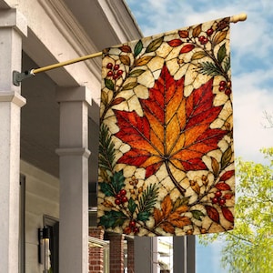 May include: A rectangular garden flag featuring a stained glass style design of a large central maple leaf in shades of red, orange, and yellow. The background has a cream-colored grid pattern with surrounding sprigs of holly, red berries, and pine.