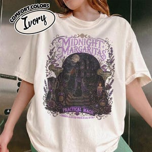 May include: An ivory t-shirt featuring a graphic print with the text Midnight Margaritas and Practical Magic. The design depicts a dark house, a full moon, a black cat, books, a blender, and margarita glasses surrounded by purple floral accents.