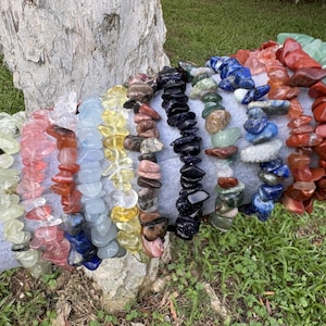 May include: A collection of eleven stretch bracelets made from various polished gemstone chips. The bracelets feature a spectrum of colors including pale green, pink, orange, light blue, yellow, brown, black, dark blue, and deep red mineral stones.