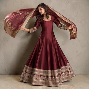 May include: A floor-length, deep maroon gown with long sleeves and a square neckline. The dress features a wide, intricate gold embroidered border at the hem and cuffs. A matching maroon dupatta with a gold border is draped overhead.