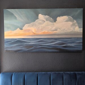 Puede incluir: Una pintura de paisaje rectangular que representa un vasto océano con olas azules profundas bajo un cielo dramático. Grandes nubes blancas y voluminosas dominan la parte superior, con tonos naranjas y dorados brillando en el horizonte.