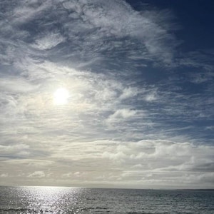 May include: A coastal scene featuring a sandy beach meeting the ocean under a bright sky. The sun shines through wispy white clouds, casting a shimmering reflection across the blue water. A gentle wave breaks near the shore in the foreground.