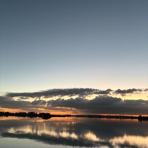 May include: A serene landscape featuring a calm body of water reflecting a sunset sky. The horizon displays a band of orange and yellow light beneath a layer of dark, horizontal clouds, transitioning into a clear, pale blue sky above.