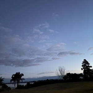 Può includere: Un paesaggio al crepuscolo con una collina erbosa in primo piano, alberi in silhouette tra cui una palma sulla sinistra, e uno specchio d'acqua calmo in lontananza sotto un vasto cielo blu punteggiato da soffici nuvole bianche sparse.