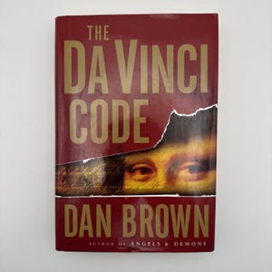 May include: A hardcover book titled The Da Vinci Code by Dan Brown. The cover is deep red with gold lettering. A torn section reveals a close-up of the Mona Lisa's eyes and handwritten script. The text Author of Angels & Demons is at the bottom.