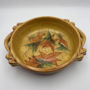 May include: A round, mustard-yellow ceramic bowl featuring two decorative handles on opposite sides. The interior base is decorated with painted orange and green maple leaf motifs and delicate vine-like line work, with small clusters of dots.
