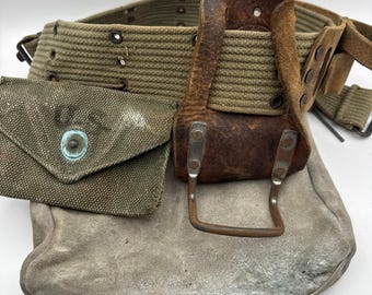 May include: An olive drab canvas military belt featuring a series of metal eyelets, a small attached canvas pouch with the letters U.S. printed on the front, a brown leather holster with a metal frame, and a large grey suede pouch attached below.