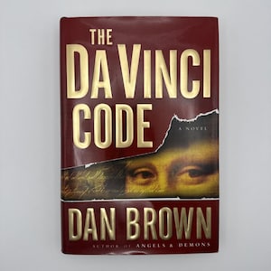 May include: A hardback book titled The Da Vinci Code by Dan Brown. The cover is deep red with gold embossed lettering. A central horizontal band features a torn paper effect revealing a close-up of the Mona Lisa's eyes and handwritten script.