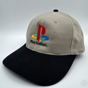Könnte beinhalten: Eine beige Baseballkappe mit schwarzem Schirm und einem schwarzen Knopf oben. Die Vorderseite zeigt ein gesticktes PlayStation-Logo in Rot, Gelb, Grün und Blau, mit dem Wort PlayStation in schwarzer Schrift direkt unter dem Logo.