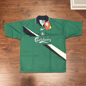 May include: A green short-sleeved jersey with a dark blue collar, featuring a white and dark blue diagonal stripe across the chest. The shirt displays the text Carlsberg, a Reebok logo, a Liverpool FC crest, and an attached red product tag.
