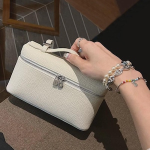 May include: A cream-colored, textured vanity case with a top handle and a silver-toned zipper closure. The bag features a structured, rectangular shape with rounded edges and a metallic zipper pull, showcasing a pebbled leather-like finish.