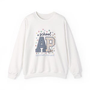 May include: A white long-sleeve sweatshirt featuring a graphic print with the text school AP assistant principal. The letters AP are large, with the A in a blue gingham pattern and the P in a floral print, accented by a small bow and colourful stars.