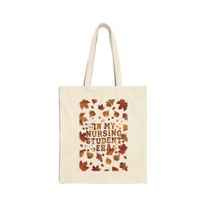 May include: A beige canvas tote bag featuring a central graphic of autumn leaves in shades of orange, red, and brown. The text "IN MY NURSING STUDENT ERA" is printed in the centre in a brown font, accompanied by small stethoscope icons.