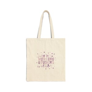May include: A beige canvas tote bag featuring a purple graphic print with the text IN MY NURSING STUDENT ERA, surrounded by small star and stethoscope icons. The bag has two long shoulder straps and a rectangular body with a flat base.