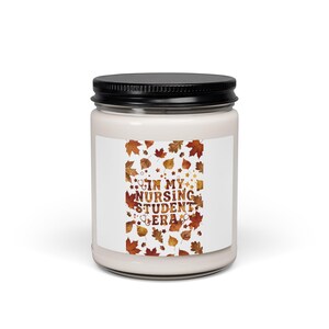 May include: A cylindrical glass candle jar with a black metal lid containing white wax. The label features a pattern of orange, red, and brown autumn leaves with the text "IN MY NURSING STUDENT ERA" printed in a bold, dark brown font.
