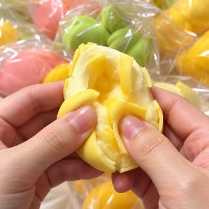 Puede incluir: Un objeto amarillo, suave y texturizado que se está separando, revelando un interior en capas. El artículo está recubierto por un material fino y flexible. Otros objetos blandos similares en rosa, verde y amarillo son visibles al fondo, cada uno envuelto individualmente en un empaque de plástico transparente.