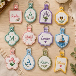 May include: A collection of twelve embroidered keychains in various shapes and colors, featuring designs such as names like Sophia, Emma, Liam, and Olivia, letters, a bee, a sailboat, a bow, lavender, and tulips, each with a metal ring attachment.