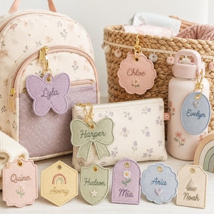 May include: A collection of personalized fabric bag tags in various shapes including butterfly, bow, scalloped, and shield designs. Each tag features embroidered names like Lyla, Chloe, Evelyn, Harper, Quinn, Avery, Hudson, Mia, Aria, and Noah with icons.
