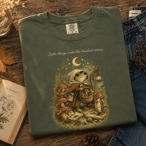 Peut inclure: Un t-shirt vert olive orné d'une illustration représentant une grenouille vêtue d'une cape à capuche, portant un panier de champignons. Le texte "Little things make the loveliest stories" est imprimé au-dessus, entouré de champignons et d'un croissant de lune.