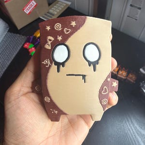 May include: A 3D-printed figure with a tan and brown color scheme. The character features two large white circular eyes with black dripping details, a straight black mouth line, and small embossed symbols including stars, hearts, and swirls.