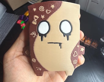 以下が含まれることがあります： ベージュと茶色の配色で3Dプリントされたフィギュア。キャラクターは、黒い滴るディテールが施された2つの大きな白い円形の目、真っ直ぐな黒い口のライン、そして星、ハート、渦巻きなどの小さなエンボス加工されたシンボルが特徴です。