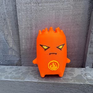 May include: A bright orange figurine with a smooth, cylindrical body and a jagged top edge. It features sharp, angular yellow eyes with black outlines, a small straight mouth, and a circular emblem on the chest displaying a stylised flame icon.