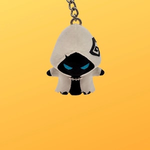May include: A keyring featuring a small, black, hooded figure with glowing blue eyes. The figure wears a white, textured cloak with a black geometric symbol on the left side of the hood. The keyring is attached to a silver-coloured metal ring.