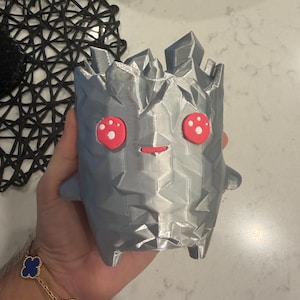 May include: A silver, low-poly, geometric-shaped planter with a faceted surface texture. The object features two bright pink circular eyes with white dots and a small horizontal pink mouth, giving it a stylized, character-like appearance.