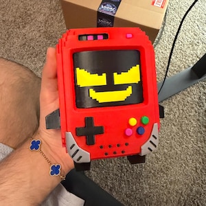 Könnte beinhalten: Eine rote, blockartige Handheld-Spielkonsole im Pixel-Art-Stil mit einem schwarzen Bildschirm, der ein gelbes, verpixeltes Gesicht zeigt. Sie verfügt über ein schwarzes Steuerkreuz, vier bunte runde Tasten und silberne Akzente an den Ecken.