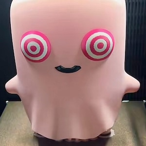 May include: A light pink, ghost-shaped figurine with a smooth, matte finish. The figure features two large, circular eyes with concentric red and white rings, and a small, black, curved mouth centred below the eyes. It has two small, pointed arms.