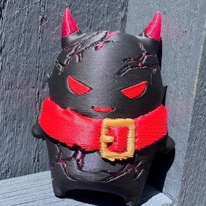 May include: A small, black, 3D-printed figurine shaped like a creature with two pointed red horns, red eyes, and a red collar featuring a tan buckle. The black body is textured with thin, recessed red lines that resemble cracks across the head and torso.