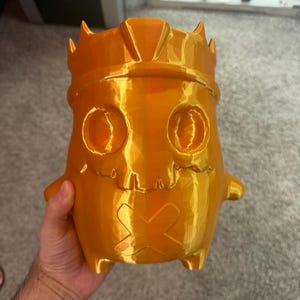 May include: A 3D printed orange container shaped like a stylised character wearing a crown. The figure features two hollow circular eye sockets, a jagged mouth area, and an X-shaped mark on the lower front section, with a smooth, glossy finish.