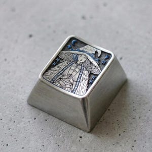 Op de afbeelding: Een zilverkleurige metalen toetsenbordtoets met een gegraveerde illustratie van een figuur met een hoed met brede rand en een textuurrijke mantel. Het ontwerp bevat blauwe accenten op de hoed, een maansikkel en kleine sterren.