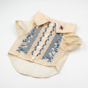 May include: A cream-colored short-sleeved button-down shirt featuring blue floral embroidery and textured gray panels along the front. The shirt includes a pointed collar and a small, triangular pin with red, blue, and yellow colors attached.