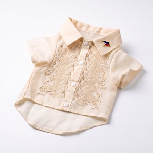 May include: A cream-colored short-sleeved button-down shirt featuring intricate tan embroidery along the front placket and a small pin shaped like the Philippine flag attached to the collar. The garment has a pointed collar and white buttons.