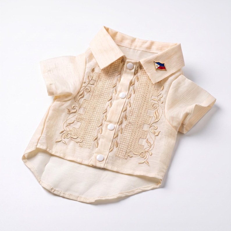 May include: A cream-colored short-sleeved button-down shirt featuring intricate tan embroidery along the front placket and a small pin shaped like the Philippine flag attached to the collar. The garment has a pointed collar and white buttons.