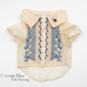 May include: A cream-colored short-sleeved polo barong shirt featuring a button-down front with white buttons. The garment is decorated with blue floral embroidery and textured panels. A small pin shaped like the Philippine flag is on the collar.