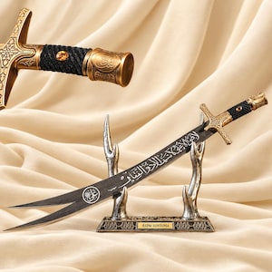 Könnte beinhalten: Ein geschwungenes Schwert mit schwarzer Klinge, weißer arabischer Kalligrafie und einem kreisförmigen Emblem. Das Heft ist goldfarben mit schwarzem Kordelgriff, einem bernsteinfarbenen Stein und einem verzierten Knauf. Es ruht auf einem silberfarbenen Geweihständer.