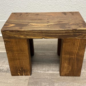 May include: A small, rectangular wooden stool featuring a dark brown stained finish that highlights the natural wood grain patterns. The stool is constructed with a flat top surface supported by four thick, sturdy legs, creating a simple, functional design.