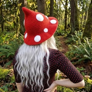May include: A red crocheted hat with a pointed top and a wide brim, featuring several circular white patches sewn onto the surface to resemble a mushroom. The hat is constructed from yarn with a visible stitch pattern throughout the fabric.