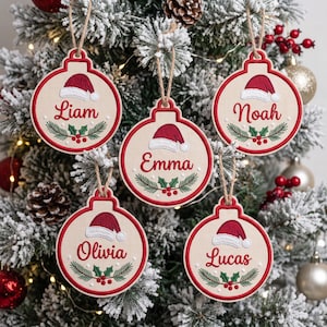 May include: Five round, cream-colored fabric ornaments with red embroidered borders and twine hangers. Each features a red Santa hat, green holly leaves with red berries, and a unique name: Liam, Emma, Noah, Olivia, or Lucas.