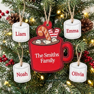May include: A large red mug-shaped Christmas ornament with the text The Smiths Family, featuring a candy cane and marshmallows. Four smaller white marshmallow-shaped ornaments hang nearby, each embroidered with the names Liam, Emma, Noah, and Olivia.