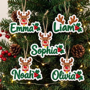 May include: Five white Christmas ornaments featuring brown reindeer with red noses, green holly leaves with red berries, and the names Emma, Liam, Sophia, Noah, and Olivia in green script, each hanging from a brown twine loop.