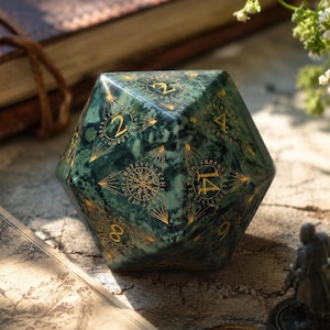 Puede incluir: Un dado de veinte caras con un patrón jaspeado verde oscuro, números grabados en color dorado y líneas geométricas intrincadas. El dado muestra números que incluyen el 2, 4, 9, 14 y 8, junto con un símbolo circular central con marcas de estilo rúnico.