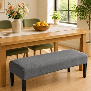 May include: A rectangular bench featuring a textured, dark grey fabric cover with a ribbed pattern. The bench is supported by four straight, black wooden legs, providing a stable base. The fabric has a consistent, woven appearance throughout.