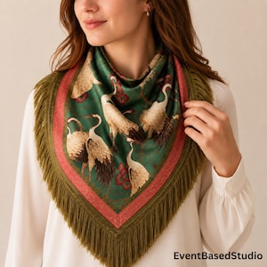 May include: A square scarf featuring a dark green background with a pattern of white cranes and red floral accents. The scarf is bordered by a wide red band and finished with long, olive green fringe along the edges. The text EventBasedStudio is visible.