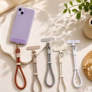 May include: A light purple smartphone attached to a brown cord wrist strap with a metal connector. Four additional wrist straps in tan, grey, cream, and white are arranged next to it, each featuring a T-shaped metal attachment point.