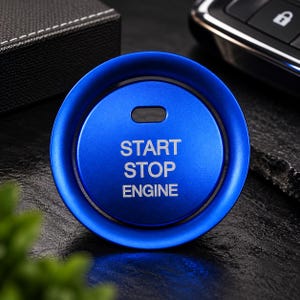 Könnte beinhalten: Eine kreisförmige blaue Abdeckung für den Motor-Start-Stopp-Knopf mit matter Oberfläche. Der Text START STOP ENGINE ist in Weiß in der Mitte aufgedruckt. Über dem Text befindet sich ein kleiner horizontaler rechteckiger Ausschnitt.