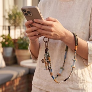 Könnte beinhalten: Ein dekoratives Handy-Armband mit einer geflochtenen Kordel in Braun-, Grün- und Blautönen, akzentuiert durch kleine stoffumwickelte Segmente, Quasten und einen Lotus-Samenkapsel-Anhänger aus Holz an einem bronzefarbenen Metallring mit Verschluss.