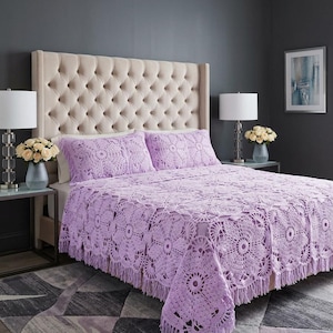 May include: A light purple crochet bedspread and two matching pillow shams featuring a repeating circular floral pattern with a fringed edge. The textile is crafted with an openwork design, showcasing intricate yarn loops and a soft lavender hue.